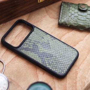 Può includere: Una custodia per telefono nera con un motivo a pelle di serpente verde e grigio. È visibile anche un porta carte abbinato in pelle di serpente verde. Gli oggetti sono su una superficie di legno, con un orologio da tasca e una scatola di legno sullo sfondo.