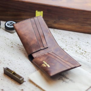 May include: Open brown leather wallet with multiple card slots and a gold logo. The wallet is displayed open, showcasing its interior design. The stitching is visible, and the wallet appears to be new. A small, off-white cloth with text is next to the wallet.