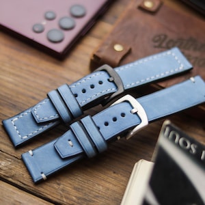 Könnte beinhalten: Zwei hellblaue Uhrenarmbänder mit weißen Nähten und Metallschnallen. Eine Schnalle ist silberfarben, die andere schwarz. Die Armbänder liegen auf einer Holzoberfläche, im Hintergrund ein Leder-Notizbuch und ein Telefon. Stilvolles Accessoire.