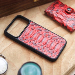 May include: A black phone case with a red snakeskin pattern is displayed on a wooden surface. A matching red snakeskin cardholder is also visible. A silver pocket watch and a green object are in the foreground.