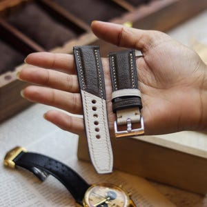 Könnte beinhalten: Zweifarbige Leder-Uhrenarmbänder in Braun und Creme mit weißen Nähten. Die Armbänder werden hochgehalten, um das Design und die Details zu zeigen. Eine Uhr mit schwarzem Armband und goldenen Akzenten ist im Vordergrund zu sehen.