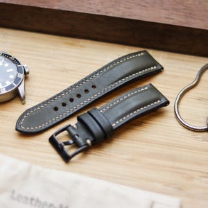 Könnte beinhalten: Ein olivgrünes Lederarmband mit weißen Nähten und einer schwarzen Schnalle wird neben einer silbernen Armbanduhr und einer silbernen Stoppuhr gezeigt. Das Armband liegt auf einer Holzoberfläche.