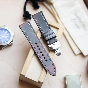 Può includere: Cinturino per orologio in due pezzi, nei toni del grigio e del bordeaux, con cuciture bianche. Il cinturino è in pelle e ha una chiusura in acciaio inossidabile. Sullo sfondo è visibile un orologio con quadrante blu. Il cinturino è esposto su un piccolo supporto di legno.