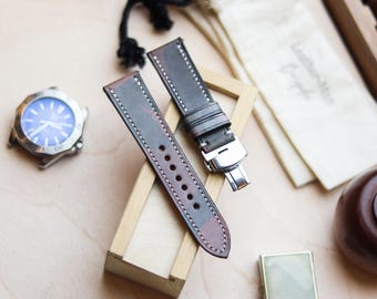 May include: Two-piece watch strap in shades of gray and burgundy, with white stitching. The strap is made of leather and has a stainless steel clasp. A watch with a blue face is visible in the background. The strap is displayed on a small wooden stand.