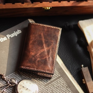 Peut inclure: Un portefeuille en cuir marron avec des coutures et des veines visibles, posé sur un journal avec le texte "war over the Punjab". Un chronomètre vintage et de petits pots de baume pour cuir sont également présents.