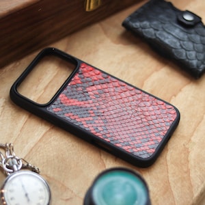 Può includere: Custodia per telefono nera con motivo a pelle di serpente rosso e grigio. La custodia è rettangolare con bordo nero. Sono visibili anche un piccolo portafoglio nero e un orologio da tasca.