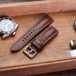 Pode incluir: Um relógio com mostrador preto e caixa prateada, pulseiras de couro castanho e um cronómetro são exibidos numa bandeja de madeira. As pulseiras do relógio têm uma superfície texturizada e uma fivela prateada. O cronómetro tem um mostrador branco.