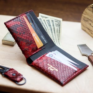 May include: A bifold wallet crafted from red and black snakeskin-textured leather, featuring multiple interior card slots containing credit cards and paper currency. A matching red snakeskin-textured leather keychain with a black metal clip is nearby.
