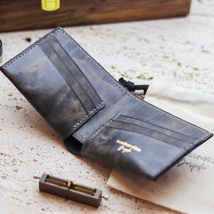 Op de afbeelding: Open, grijze leren portemonnee met kaartsleuven. De portemonnee heeft een distressed afwerking en is gestikt met witte draad. De woorden "Leather-Man Bespoke" staan op een kleine tas.