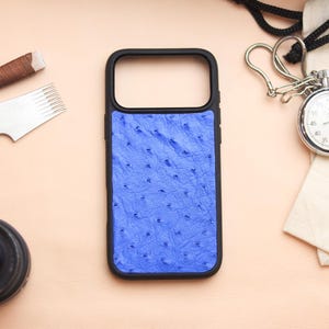 Può includere: Una custodia per telefono nera con un design blu strutturato. La custodia è circondata da un obiettivo della fotocamera, un cronometro e strumenti per la lavorazione della pelle. Il cronometro ha un cordino nero e un quadrante bianco.