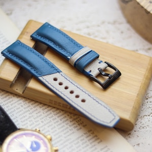 Può includere: Cinturini per orologi in pelle blu e bianca con fibbia nera, esposti su un blocco di legno. I cinturini hanno uno strato superiore blu chiaro e un lato inferiore bianco con cuciture. I cinturini dell'orologio sono appoggiati su un blocco di legno.
