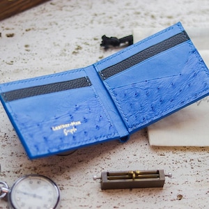 May include: A bright blue bifold wallet featuring a textured ostrich leather finish with smooth black leather card slots. The interior is stamped with the gold text Leather-Man Bespoke. The wallet is open, displaying multiple compartments.