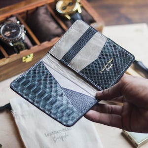 Puede incluir: Una cartera plegable hecha de cuero azul oscuro con textura de piel de serpiente y cuero liso gris claro. El interior cuenta con varias ranuras para tarjetas y el texto dorado Leather-Man Bespoke en el lado derecho, con costuras detalladas.