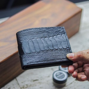 May include: A black bifold wallet featuring a textured ostrich skin pattern on the exterior, accented by a smooth, raised central panel with a series of horizontal, rounded ridges running across the front surface of the folded accessory.