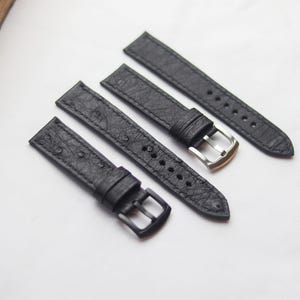 Könnte beinhalten: Vier schwarze Uhrenarmbänder mit strukturierten Oberflächen und Metallschnallen. Zwei Armbänder haben silberne Schnallen und zwei haben schwarze Schnallen. Die Armbänder sind auf einer weißen Oberfläche angeordnet.