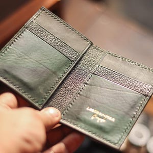 May include: Open, dark green leather wallet with multiple card slots. The wallet has a textured, stitched design and the words "Leather Man Forge" are embossed in gold lettering.