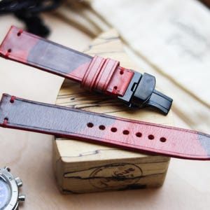 Può includere: Cinturini per orologi in pelle rossa e nera con chiusura nera. I cinturini sono esposti su un blocco di legno. I cinturini hanno cuciture rosse e un motivo a colori sfumati.