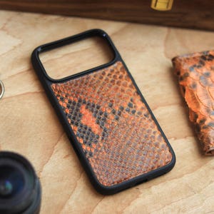May include: A black phone case featuring an orange and brown snakeskin design. A small wallet with a similar pattern is also present. The items are displayed on a wooden surface, alongside a pocket watch and a camera lens.