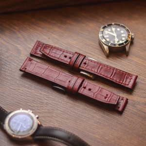 Könnte beinhalten: Burgunderfarbene Lederarmbänder mit Krokodilmuster liegen auf einer Holzoberfläche. Eine Vintage-Uhr mit schwarzem Zifferblatt und goldfarbenem Gehäuse befindet sich in der Nähe, zusammen mit einer zweiten Uhr und Garnrollen.