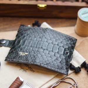 Peut inclure: Un portefeuille en cuir texturé gris foncé avec un motif d'écailles, une fermeture à bouton-pression et le texte "Leather Man Design". Le portefeuille est sur un tissu beige avec d'autres accessoires.