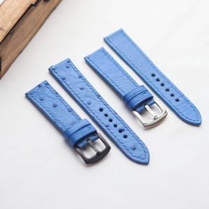 Könnte beinhalten: Vier blaue Uhrenarmbänder mit strukturierter Oberfläche vor weißem Hintergrund. Zwei Armbänder haben silberne Schnallen, zwei schwarze. Eine Holzkiste ist teilweise oben links zu sehen.