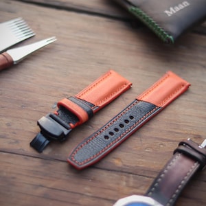 Könnte beinhalten: Zweifarbige Uhrenarmbänder aus orangefarbenem und schwarzem Leder mit roten Nähten. Die Armbänder werden auf einer Holzoberfläche zusammen mit Uhrmacherwerkzeugen und einer Ledergeldbörse mit dem eingeprägten Namen "Maan" präsentiert.