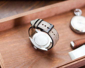 May include: A wristwatch with a light gray textured leather strap and a black metal buckle. The watch case has a silver-toned back and is positioned to show the strap's underside and the buckle mechanism clearly.