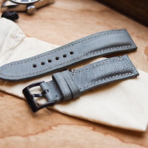 Könnte beinhalten: Zwei graue Leder-Uhrenarmbänder mit schwarzen Schnallen, die auf einem kleinen, cremefarbenen Stoffbeutel liegen. Die Armbänder sind strukturiert und genäht, mit mehreren Löchern zur Anpassung. Die Armbänder liegen auf einer Holzoberfläche.