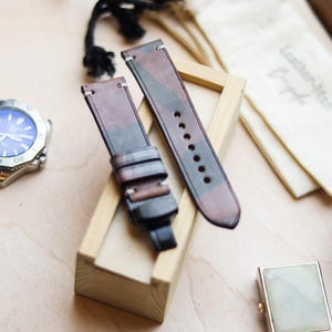 Può includere: Cinturino per orologio in due pezzi con motivo mimetico marrone, verde e grigio. Il cinturino è in pelle e ha una chiusura con fibbia. Sullo sfondo è visibile un orologio argentato con quadrante blu.