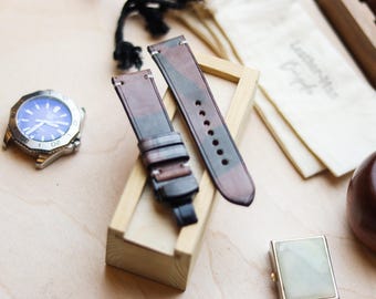 May include: Two-piece watch strap in a camouflage pattern of brown, green, and gray. The strap is made of leather and has a buckle closure. A silver watch with a blue face is visible in the background.