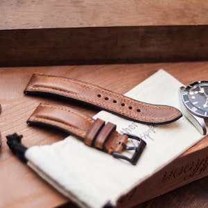 Pode incluir: Uma pulseira de relógio de couro marrom com fivela preta e um relógio prateado com mostrador preto. O relógio e a pulseira estão sobre uma superfície de madeira com uma pequena bolsa e um relógio de bolso.