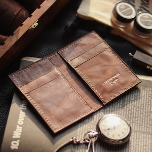 May include: An open, brown leather wallet with card slots, stamped with "Leather-man" in gold lettering. A vintage pocket watch and other accessories rest on a newspaper. The wallet is on a dark surface.