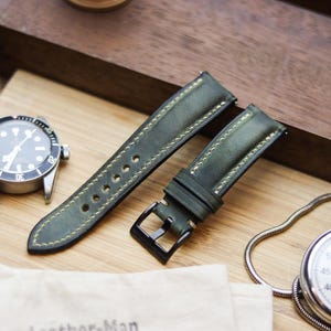 Könnte beinhalten: Grünes Leder-Uhrenarmband mit cremefarbenen Nähten und schwarzer Schnalle. Eine silberne Uhr mit schwarzem Zifferblatt und eine Stoppuhr sind ebenfalls auf einer Holzoberfläche zu sehen. Das Uhrenarmband ist etwa 20 cm lang.