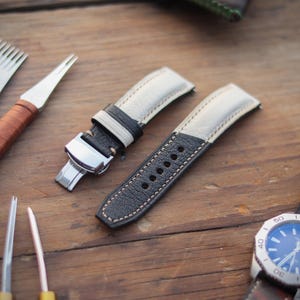 Könnte beinhalten: Zwei schwarz-weiße Lederarmbänder mit silberner Schließe, Uhrmacherwerkzeuge und eine Armbanduhr mit blauem Zifferblatt und braunem Lederarmband. Die Artikel sind auf einer Holzoberfläche angeordnet.