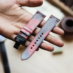 Può includere: Un paio di cinturini per orologi con un motivo mimetico rosso e grigio. I cinturini sono in pelle e hanno cuciture rosse. I cinturini sono mostrati su uno sfondo sfocato.