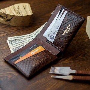 May include: A brown bifold wallet with a textured snakeskin-patterned exterior and smooth interior leather. The wallet features multiple card slots containing various credit cards and a compartment holding several US dollar bills. The text Leather-Man is embossed in gold on the inner flap.