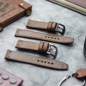 Könnte beinhalten: Braune Leder-Armbänder mit silbernen und schwarzen Schnallen, auf einer grauen Oberfläche. Ein Leder-Notizbuch und ein Kameraobjektiv sind ebenfalls zu sehen. Die Armbänder sind neu.