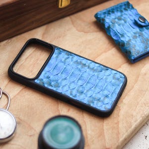 May include: A black phone case with a blue snakeskin pattern rests on a wooden surface, alongside a matching wallet. A silver pocket watch and a wooden box are also present. The phone case and wallet have a textured, scaly appearance.