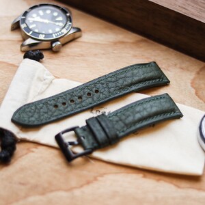 Può includere: Cinturini per orologi in pelle verde con superficie strutturata, esposti su un sacchetto color crema. Sono visibili anche un orologio argentato e un piccolo timer argentato. I cinturini hanno una fibbia in metallo scuro.