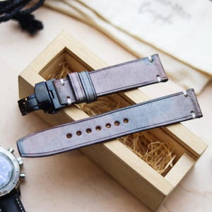 Può includere: Cinturini per orologi in pelle marrone con cuciture bianche e chiusura nera, esposti in una scatola di legno con paglia. Un orologio nero e argento è visibile a sinistra. I cinturini hanno un effetto sfumato.