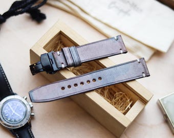 May include: Brown leather watch straps with white stitching and a black clasp, displayed in a wooden box with straw. A black and silver watch is visible on the left. The straps have a gradient color effect.
