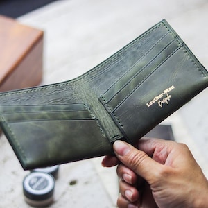 Könnte beinhalten: Geöffnetes, olivgrünes Lederportemonnaie mit mehreren Kartenfächern. Das Portemonnaie hat ein Used-Finish und den Text "Leather-Man Design" in goldfarbenen Buchstaben. Es wird offen gehalten und zeigt sein Inneres.