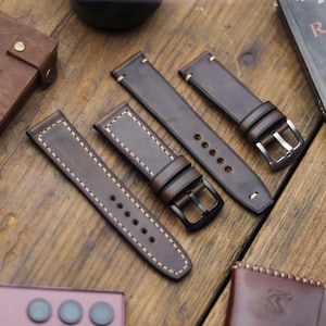 Può includere: Quattro cinturini per orologi in pelle marrone con cuciture bianche e fibbie in metallo sono disposti su una superficie di legno. I cinturini variano nel design, alcuni con fori per la regolazione e altri con un aspetto semplice e pulito. La pelle ha un aspetto invecchiato.