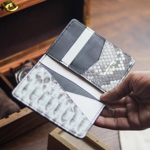 Op de afbeelding: Een opvouwbare kaarthouder met een combinatie van zwart leer en grijs en wit leer met slangenprint. De binnenzijde bevat meerdere kaartsleuven met witte en zwarte lederen accenten en zichtbare stiksels langs de randen.