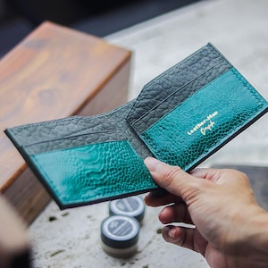 May include: Open leather wallet with a textured, dark gray exterior and teal interior. The teal sections have a reptile-like pattern. The wallet is open, revealing card slots and the gold text "Leather-Man Graph".