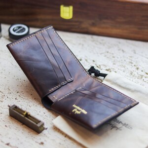 May include: Open brown leather wallet with multiple card slots and a gold logo. The wallet is displayed open on a light-coloured surface, with a small canvas bag and a metal tool nearby. The stitching is a light tan colour.