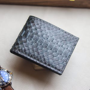 May include: A grey bi-fold wallet with a textured snakeskin pattern. The wallet is made of leather and has a black border. A brown leather watch with a black and gold watch face is next to the wallet.