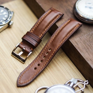 Pode incluir: Close-up de duas pulseiras de relógio de couro castanho com costuras contrastantes e uma fivela prateada. Um relógio prateado e um cronómetro vintage também são visíveis, sugerindo um tema de cronometragem e acessórios.