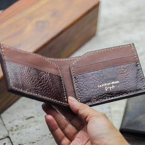 May include: A bifold wallet crafted from dark brown leather with a textured, pebbled finish. The interior features smooth brown leather card slots and a central bill compartment. The text "Leather-Man Bespoke" is embossed in gold on the right side.