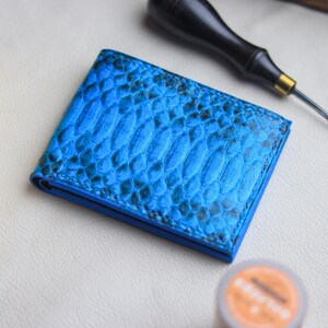 May include: A vibrant blue bifold wallet featuring a textured snakeskin pattern with dark grey scale details. The wallet is constructed with visible stitching along the edges and has a slim, rectangular profile with a smooth blue interior lining.